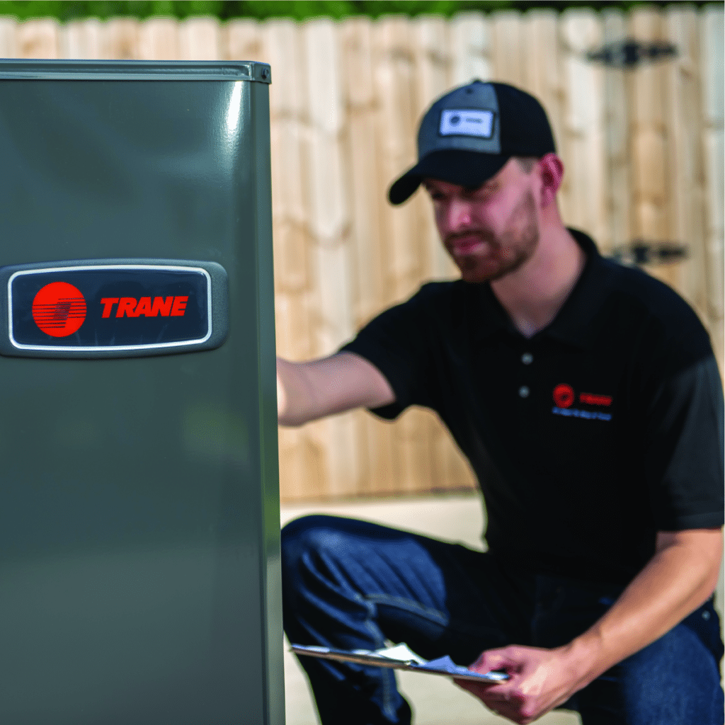 an AC technician kneeling down to perform AC maintenance on a Trane system